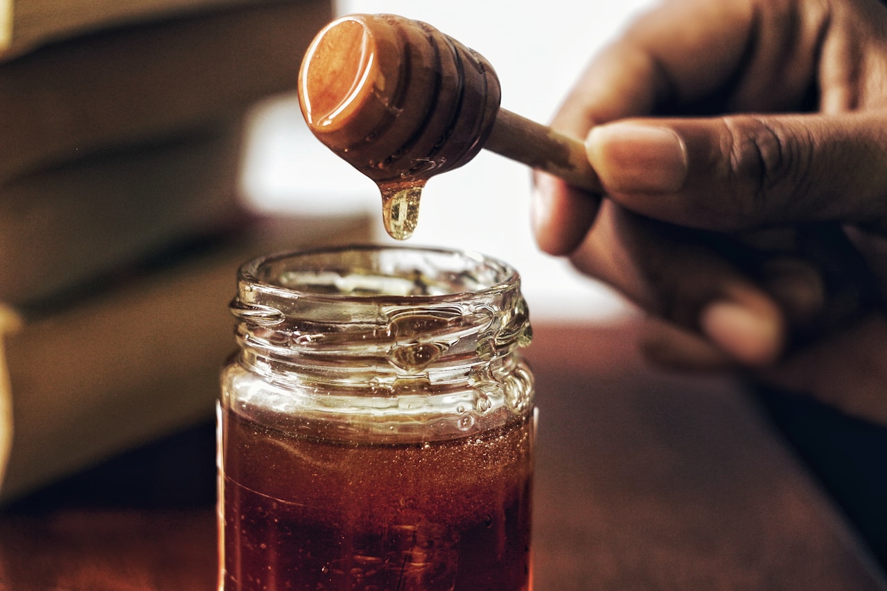 Wooden honey dipper with a thick drop of amber liquid honey falling into a glass jar
