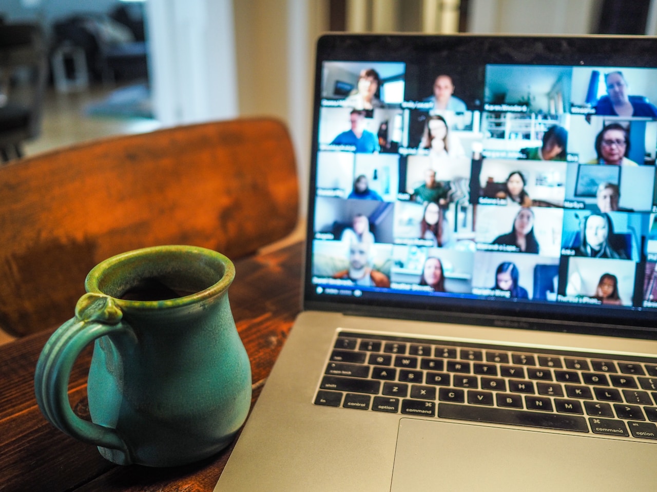 Laptop on a wooden table showing a video conference with many participants and a mug nearby